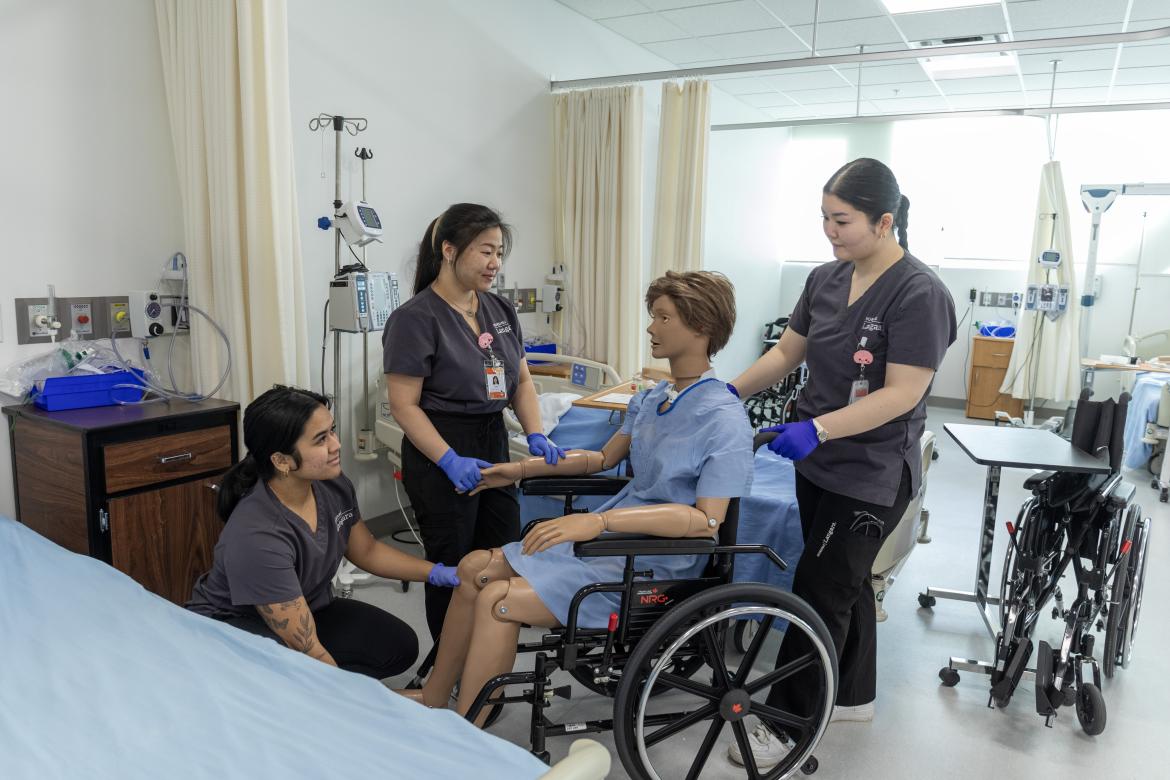 A trio of nursing students train how to handle a patient in a wheelchair using a nursing doll.