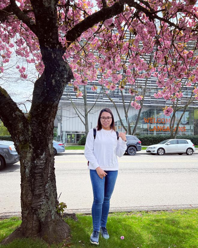 Hnin (Elisa) Pwint under a cherry blossom tree across the street from Langara College with the Science and Technology Building in the background.
