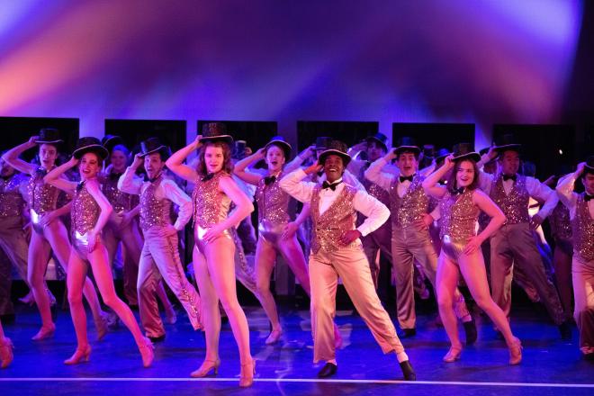 Dancers on a stage dressed in shiny gold bodysuits and shiny gold vests hold their top hats. The lighting is purply pink.
