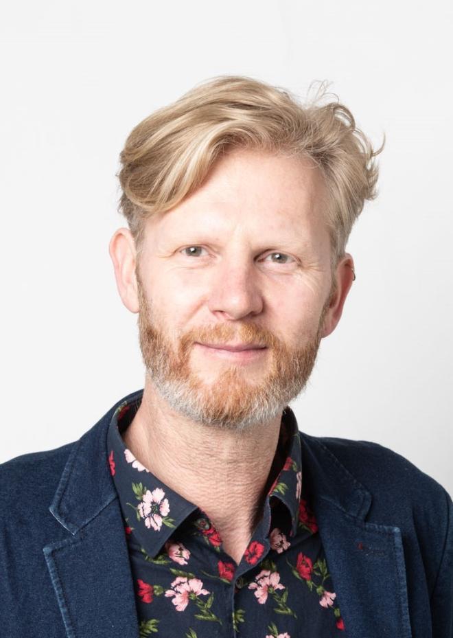 Amiel Gladstone: blonde hair quaffed to the side. A red beard with flecks of grey. A floral pattern shirt below a navy sportcoat.