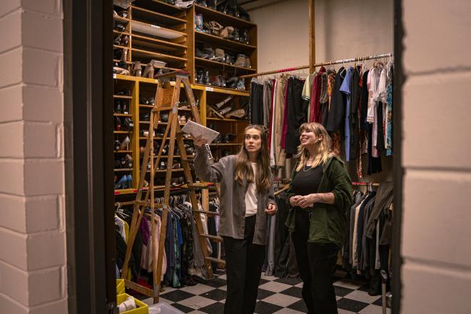 Two students stand in a costume room filled with clothes. One of them points upwards with a notebook.