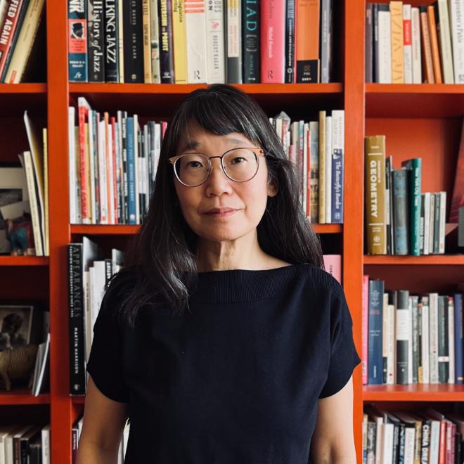 Madeleine Thien portrait standing in front of an orange bookshelf filled with literary works. Photo by Rawi Hage.