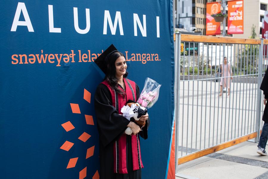 A Langara grad poses in front of an "Alumni" banner at convocation.