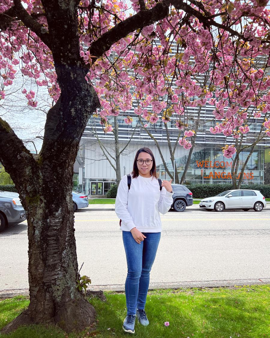 Hnin (Elisa) Pwint under a cherry blossom tree across the street from Langara College with the Science and Technology Building in the background.