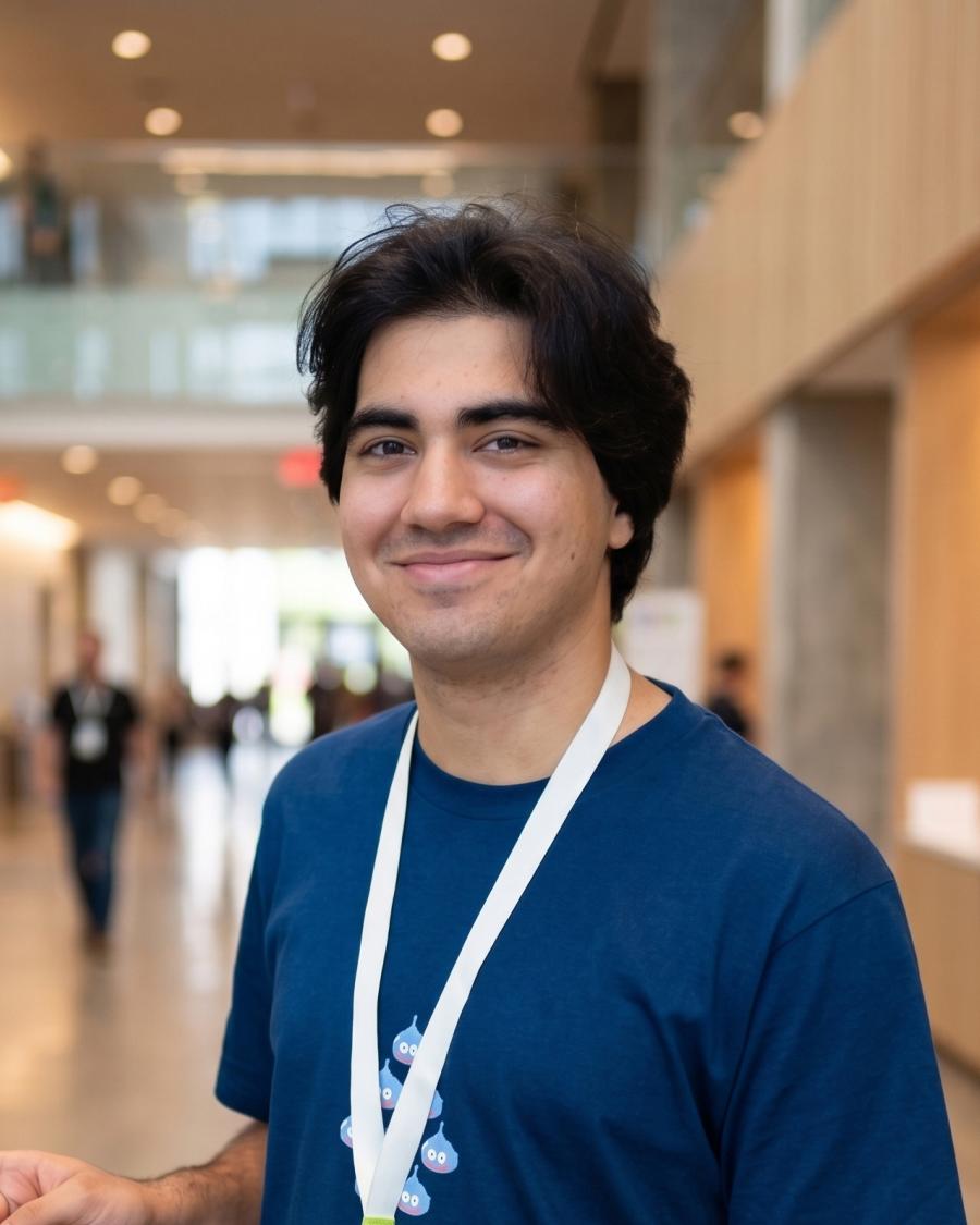 Smiling student with dark, medium-length hair wearing a blue T-shirt and a white lanyard, standing in a bright, modern hallway with people and glass walls in the background.
