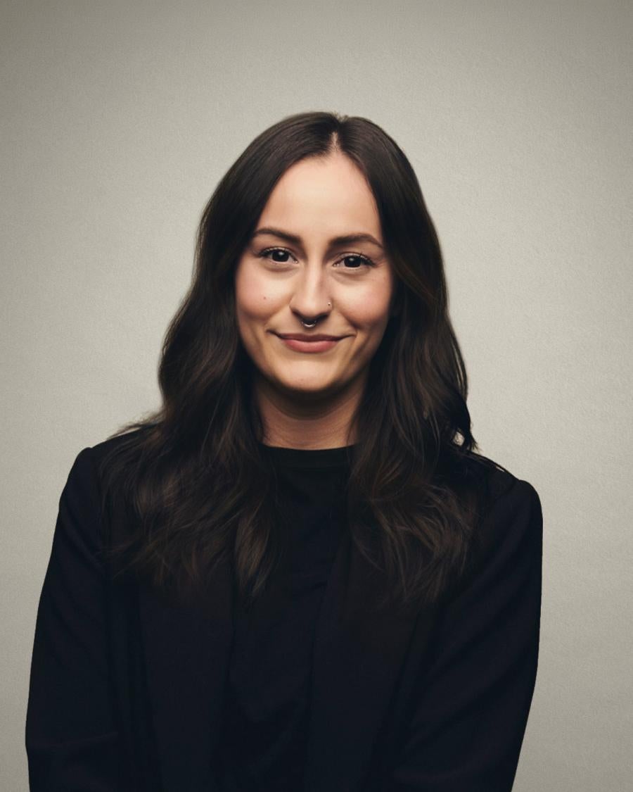 Smiling person with long dark wavy hair and a nose ring, wearing a black top and blazer, posed against a plain light-grey background.