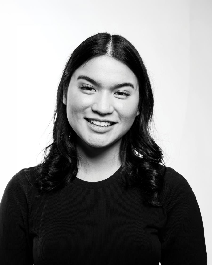 Smiling portrait of a person with shoulder-length dark hair wearing a dark top, shown in black and white against a plain light background.