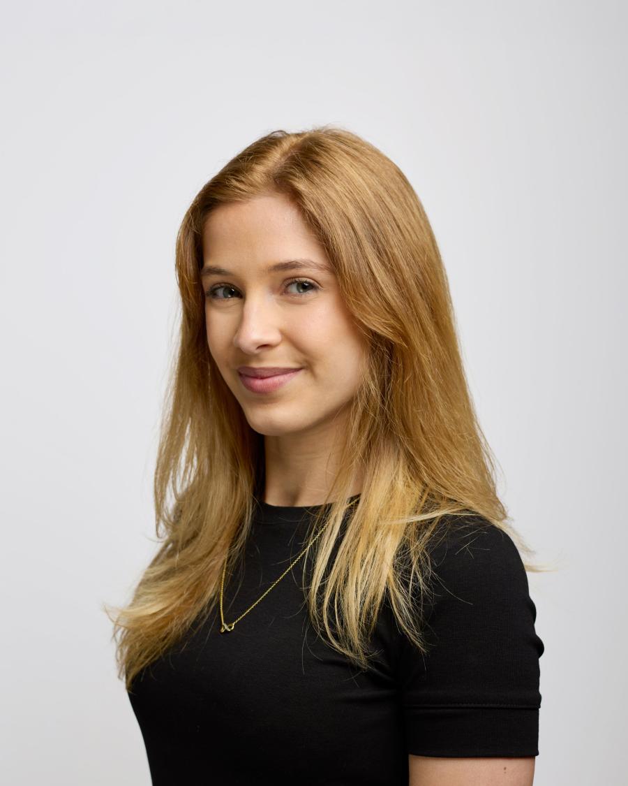 Smiling person with long light brown hair wearing a black top and a delicate necklace, shown in a side-facing portrait against a plain light background.