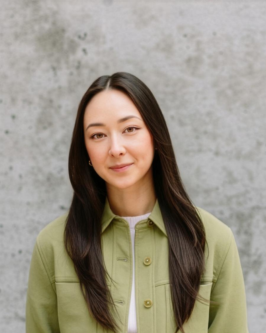 Person with long straight hair wearing a light green jacket and black jeans, standing against a textured gray concrete wall.