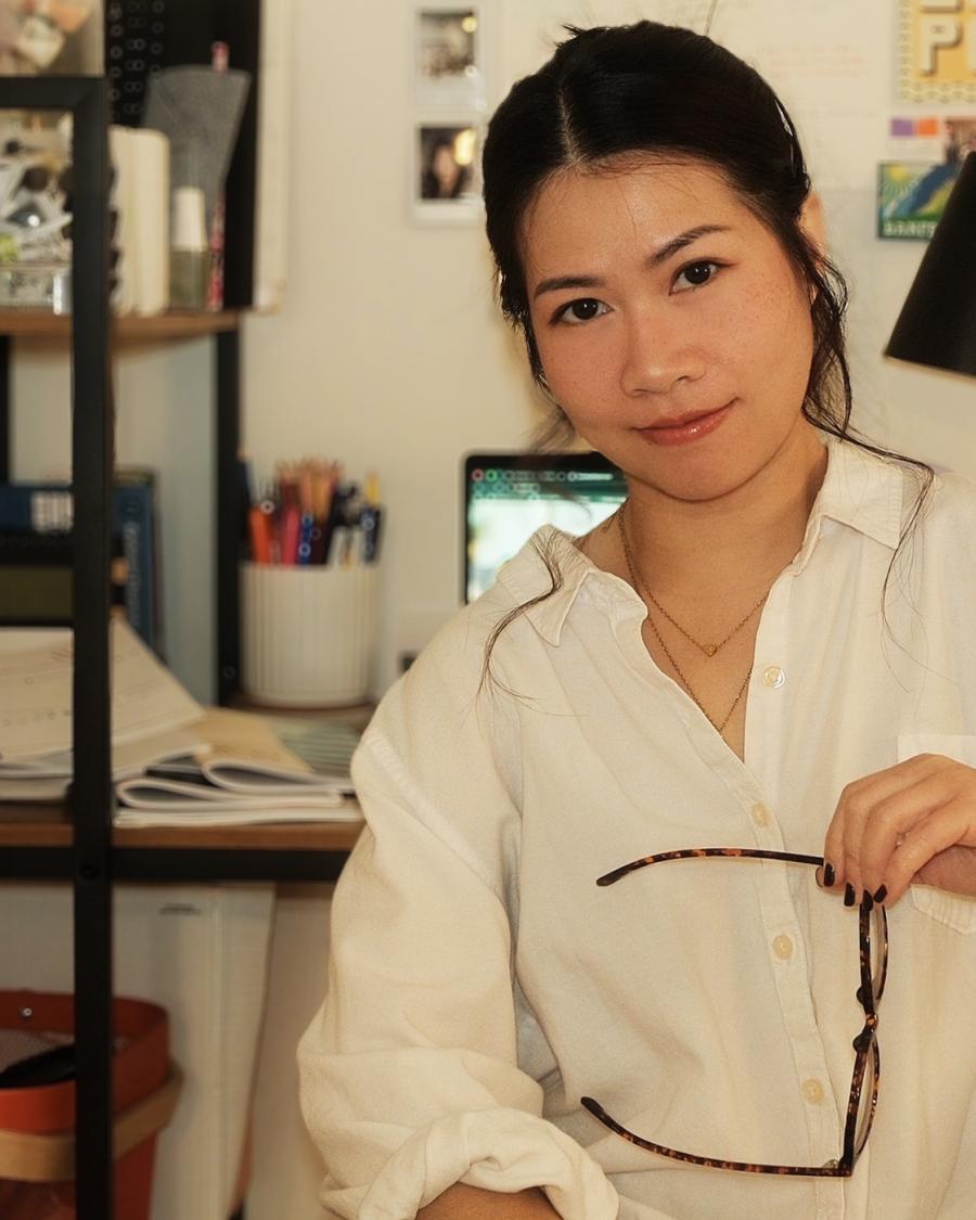 Person sitting in a home office, holding glasses and looking at the camera, with a desk, laptop, books, and photos visible in the background.