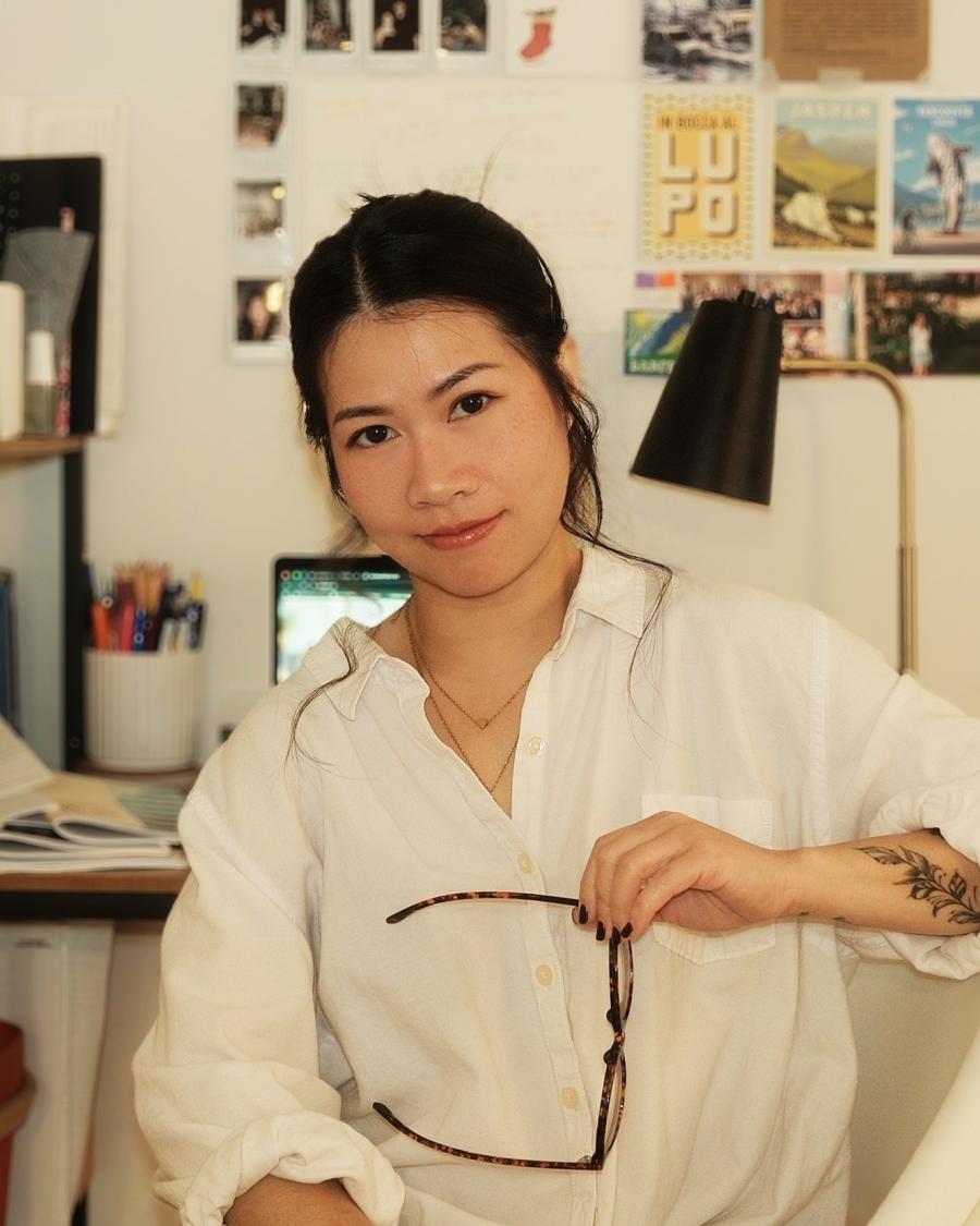Smiling student wearing a white button-up shirt, holding a pair of glasses while seated in a creative workspace with a desk, lamp, and photo-covered wall in the background.
