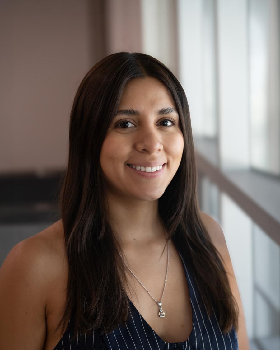 Smiling student with long dark hair wearing a navy pinstriped sleeveless top and a silver necklace, standing indoors beside large windows with a softly blurred background.