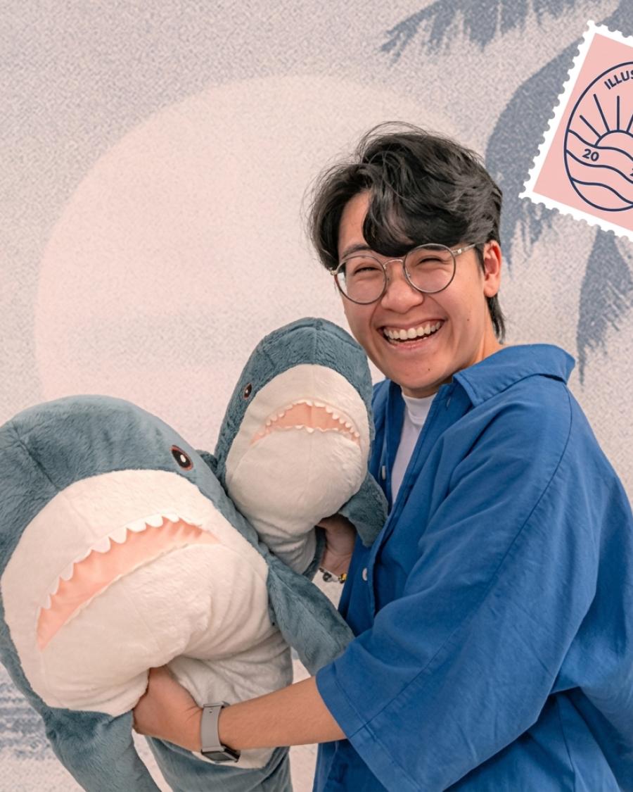 Smiling person wearing glasses and a blue shirt, holding two large plush shark toys in front of a pastel illustration-style background with a small stamp graphic that reads “Illustration 2023.”