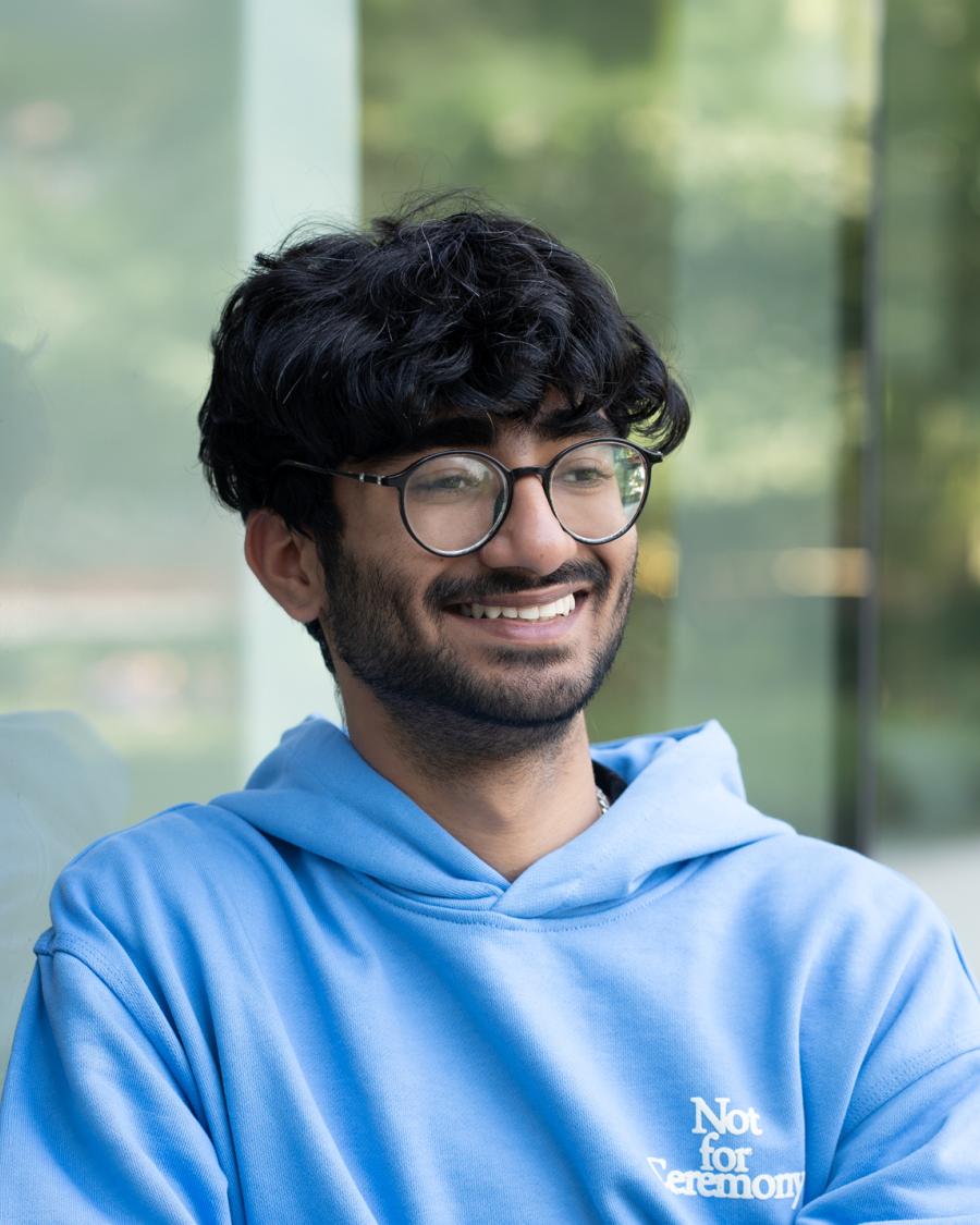 Smiling student wearing round glasses and a light blue hoodie, standing outdoors beside a glass wall with a blurred green background.