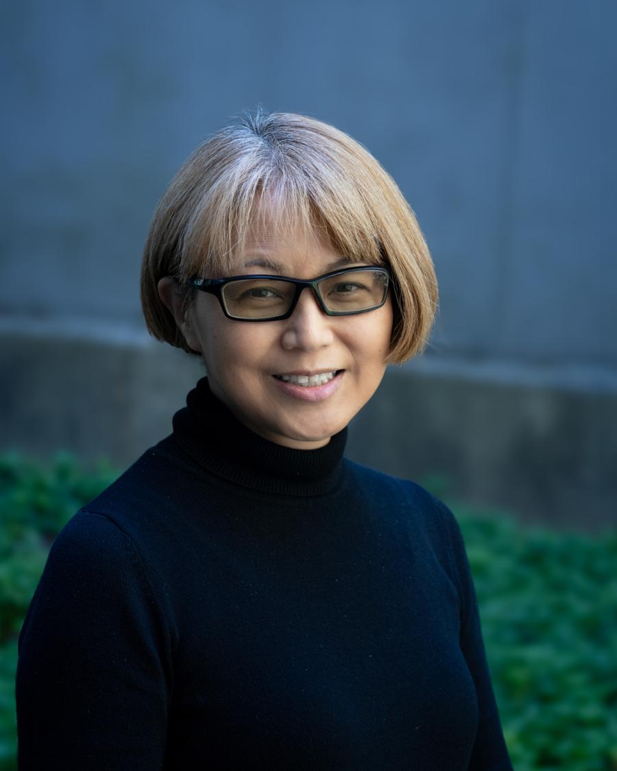 Smiling person with short light brown hair and glasses, wearing a black turtleneck, standing outdoors in front of a blurred blue wall and green foliage.