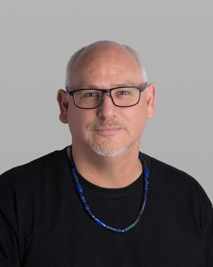 Headshot of Aaron Nelson, with a short grey beard, dark rectangular glasses, a black T-shirt and a blue beaded necklace, against a grey background.