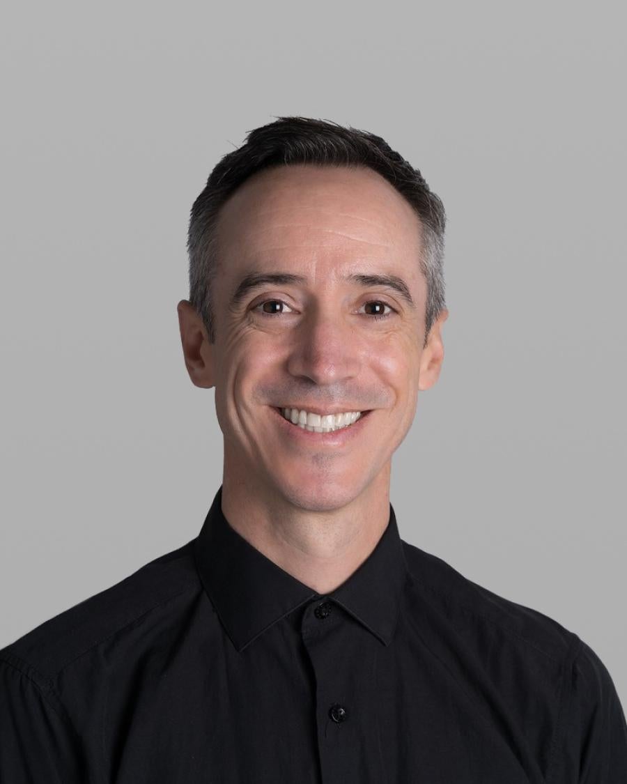 Headshot of Andres Lara Seedorf, smiling, with short greying hair and a black button-up shirt, against a grey background.