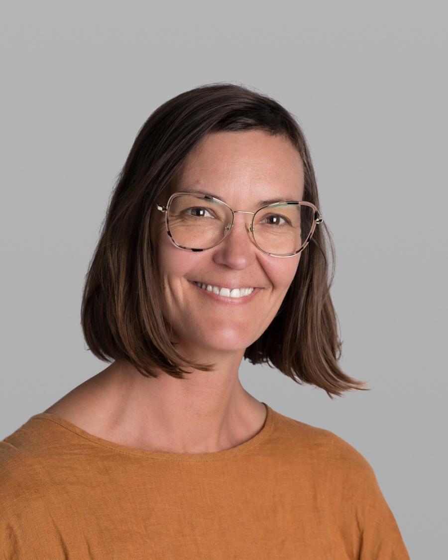 Headshot of Claire Wood, smiling, with shoulder-length brown hair, gold wire-rimmed glasses and a rust-orange sweater, against a grey background.