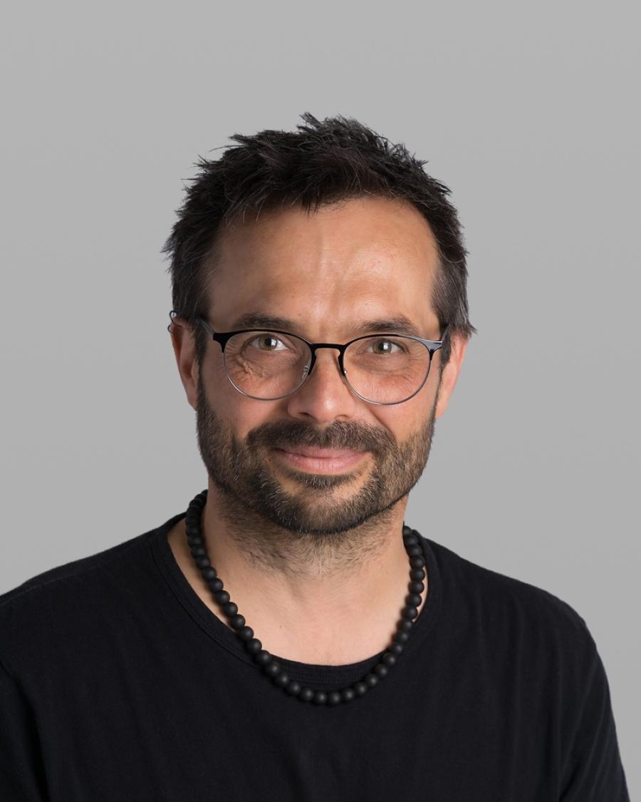 Headshot of Dave Savard, with dark hair, a dark beard, round black glasses, a black T-shirt and a beaded necklace, against a grey background.