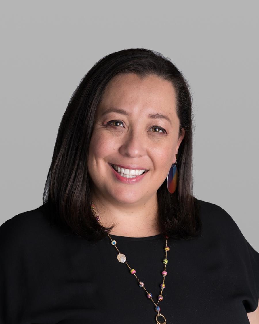 Headshot of Jessica Acevedo Castaneda, smiling, with long dark hair, colourful earrings, a black top and a long beaded necklace, against a grey background.