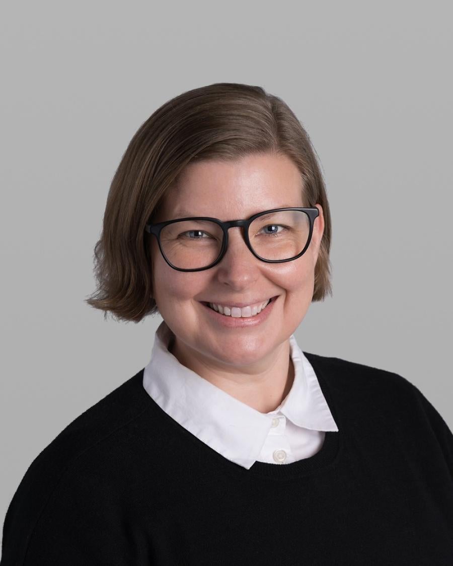 Headshot of Katherine Davis, smiling, with a light brown bob, black glasses and a black sweater over a white collared shirt, against a grey background.