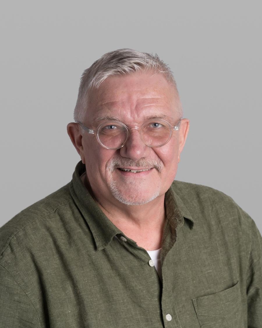 Headshot of Kevin Smith, smiling, with white hair, a short white beard, thin round glasses and an olive-green shirt, against a grey background.