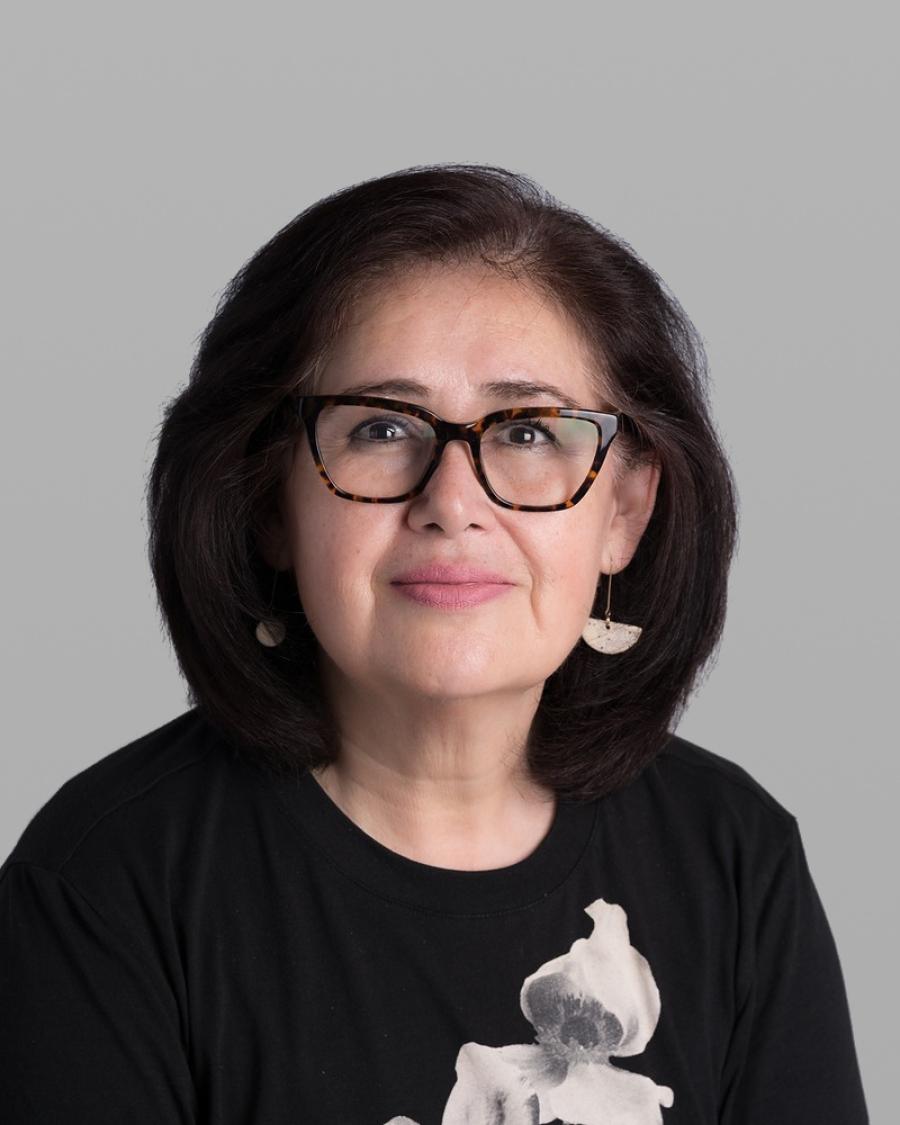 Headshot of Marcela Noriega, with dark shoulder-length hair, bold black glasses and a black T-shirt with a white print, against a grey background.