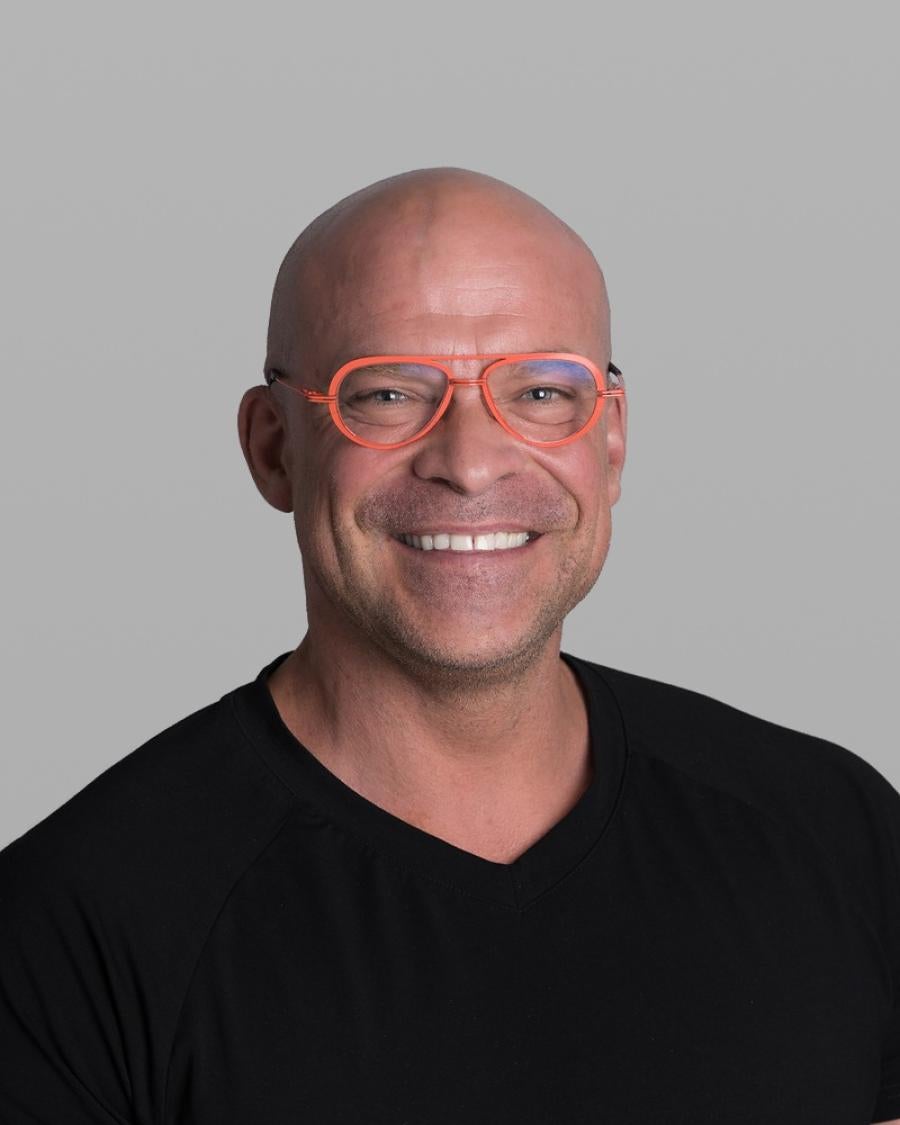 Headshot of Peter Vysek, smiling broadly, with a shaved head, coral-orange glasses and a black V-neck T-shirt, against a grey background.