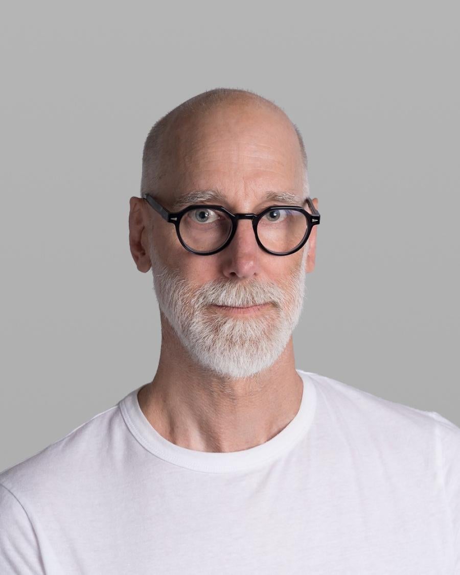Headshot of Scot Geib, with a shaved head, a white beard, round black glasses and a white T-shirt, against a grey background.