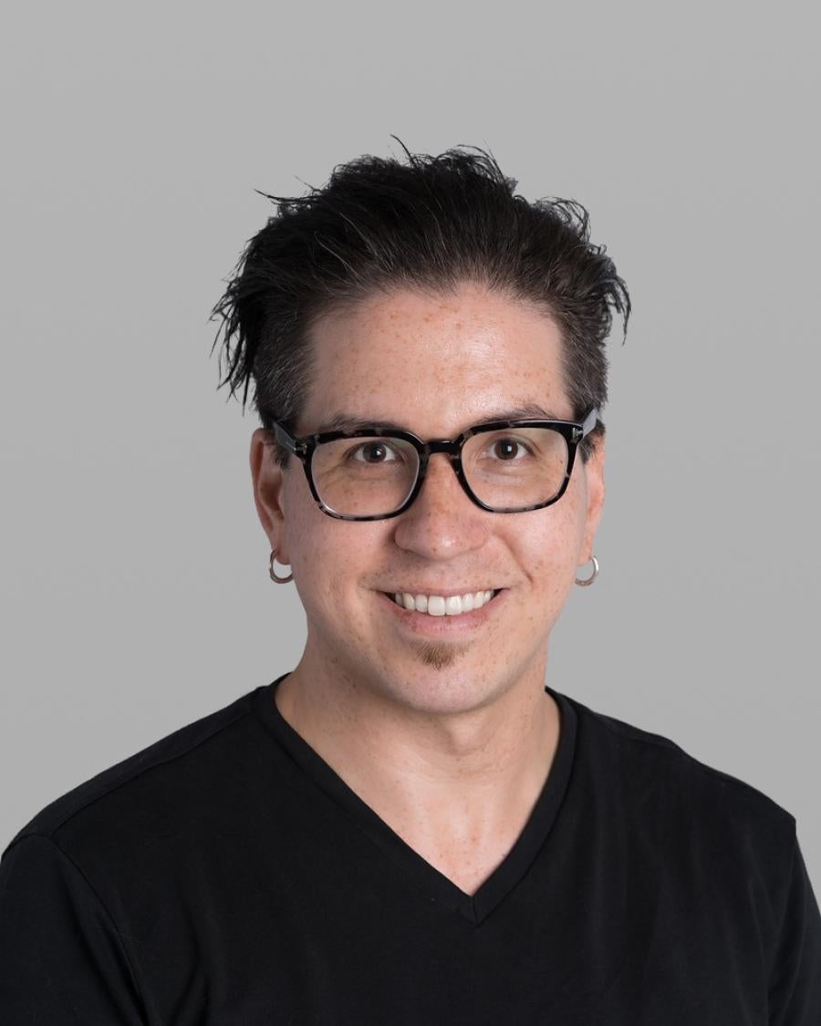 Headshot of Tyler Higgs, smiling, with dark tousled hair, black-framed glasses, small hoop earrings and a black V-neck T-shirt, against a grey background.