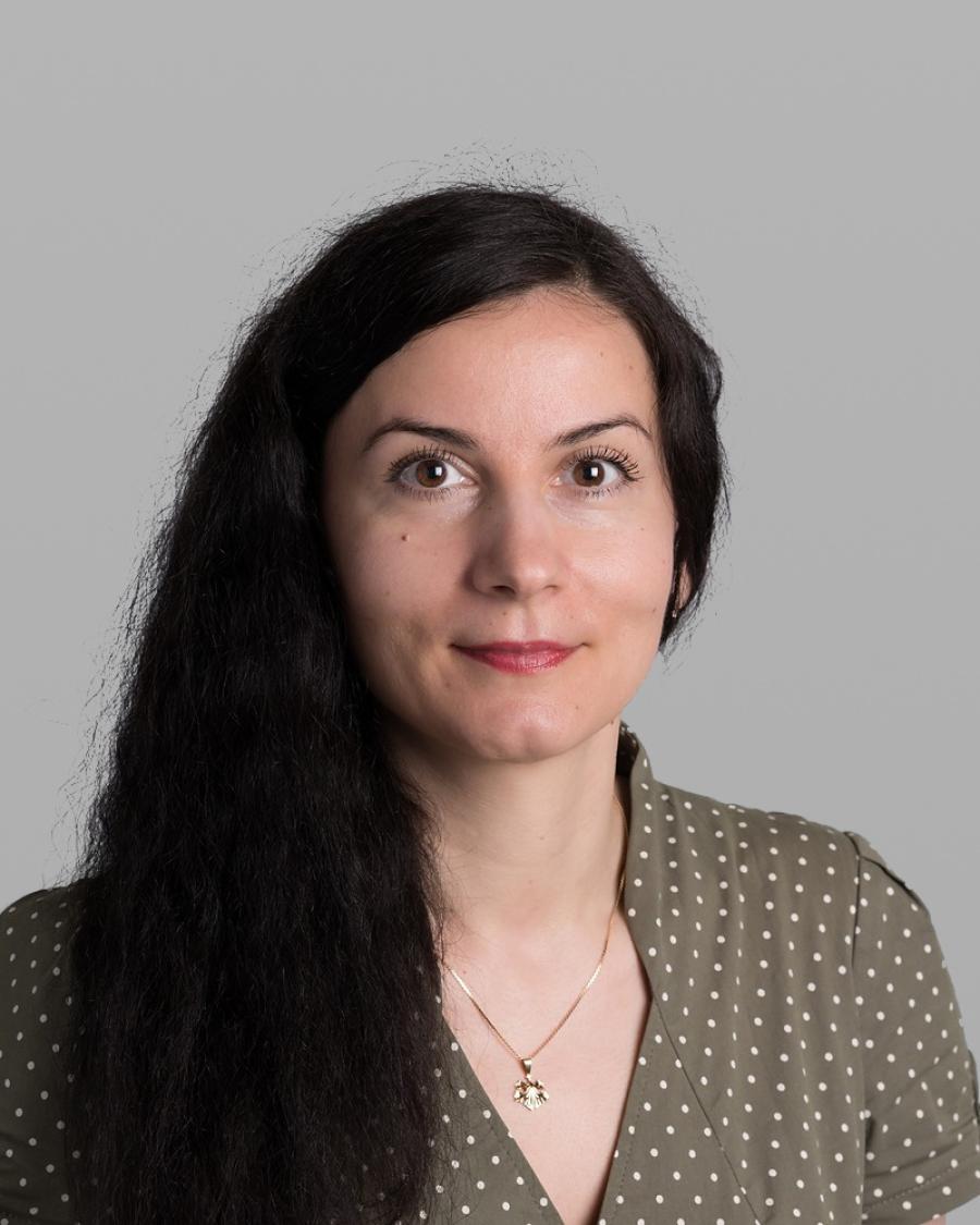 Headshot of Valentina Abanina, with long dark wavy hair, a small pendant necklace and an olive-green polka-dot blouse, against a grey background.