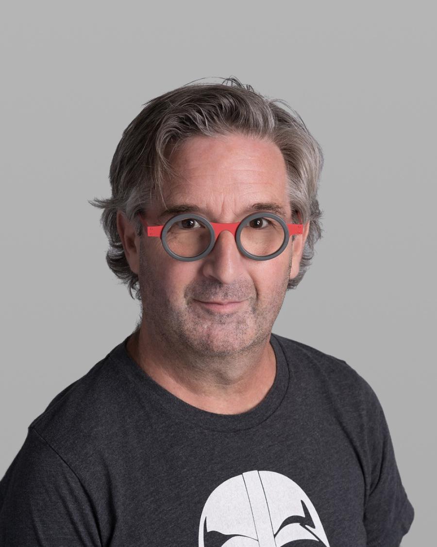 Headshot of Yvan Morissette, with wavy grey hair, round red-and-grey glasses and a dark grey printed T-shirt, against a grey background.